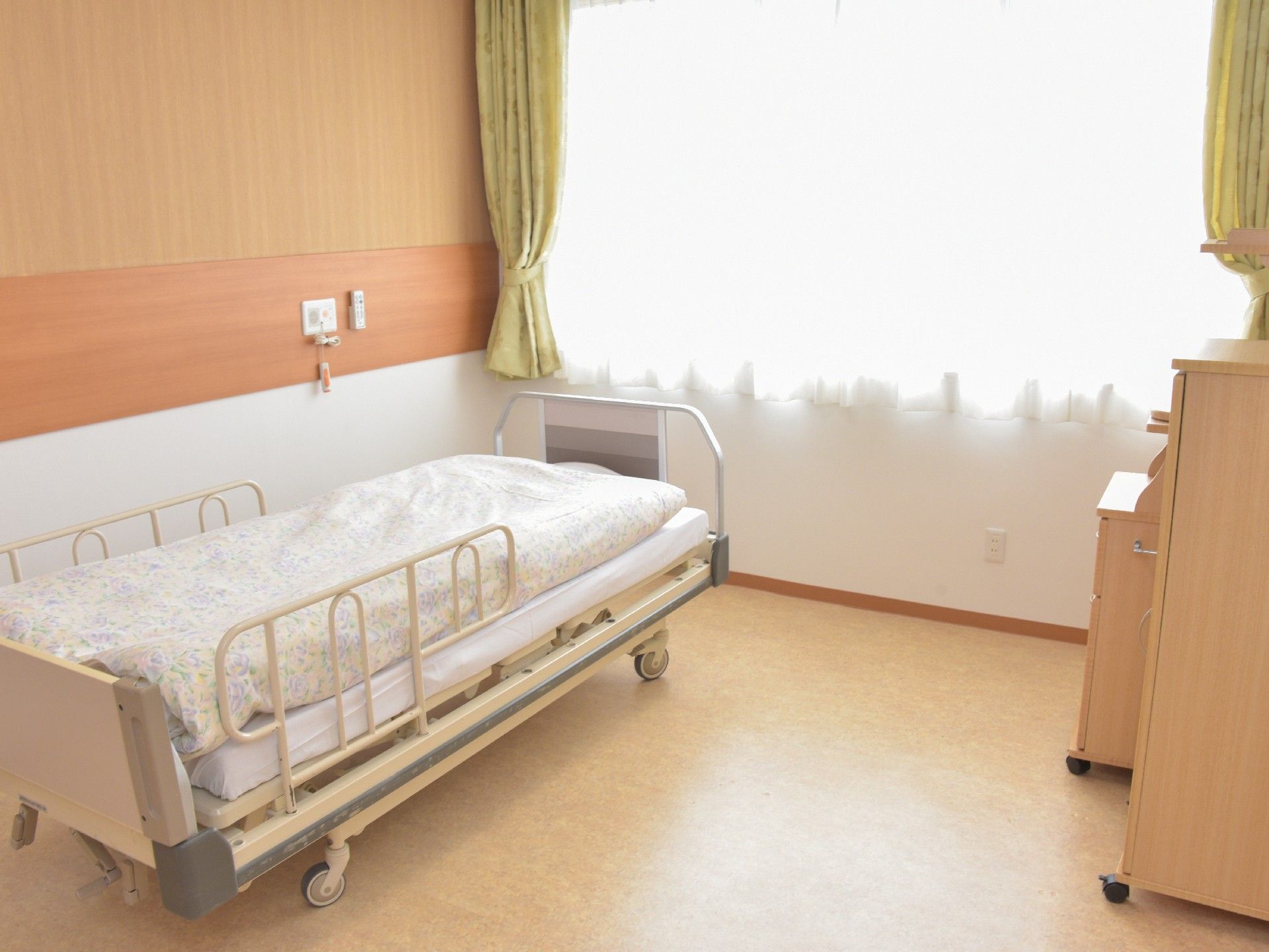 居室の壁には大きな腰高窓があり、レースと厚地のカーテンが掛けられている。壁際に置かれたベッドにはフェンスが付いている。
