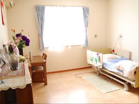 居室はゆったりとした広さがあり、右側にはシングルベッド、左側には机が配置されている。奥の窓には、青色のカーテンが掛けられている。