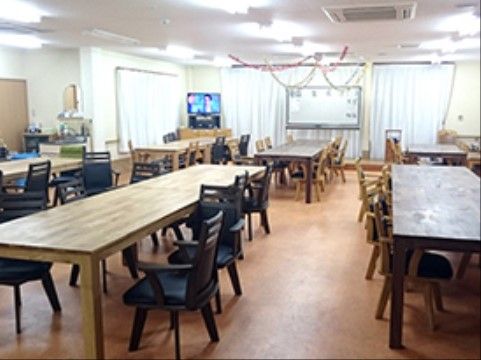 広々としたリビングには、木目調のテーブルと黒い椅子がたくさん並べられている。部屋の奥には、テレビとホワイトボードが置かれている。