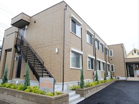 外階段が設けられた施設である。タイル貼りの外壁で、建物周りには緑が植えられている。銀色の看板に施設名がオレンジ色で表示されている。