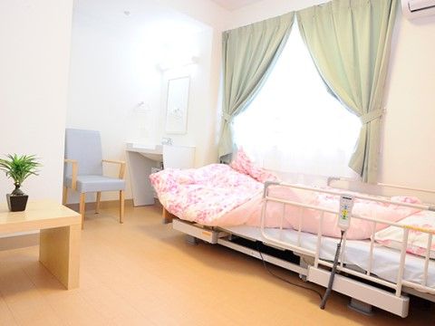 緑色のカーテンをかけた窓の側に、ピンク色の花柄が入った掛け布団カバーが特徴のベッドを置いている。奥に洗面台を設けている。