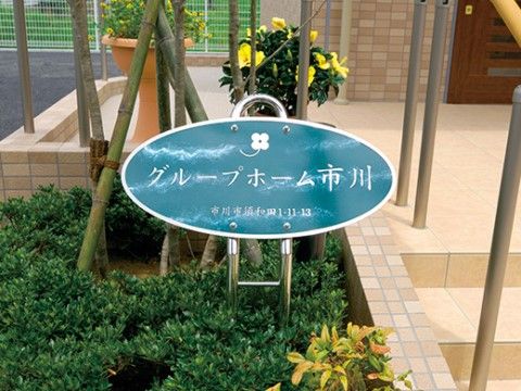 階段脇の緑の生い茂る植え込みに、施設のサインボードが設置されている。住所と施設名の書かれた楕円形の看板は、洗練されたデザインである。