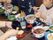 入居者様達が食事をしている様子である。みんなで座卓に座り、そば、天丼、漬物のセットメニューを食していて、スタッフが天かすを入れてあげている。