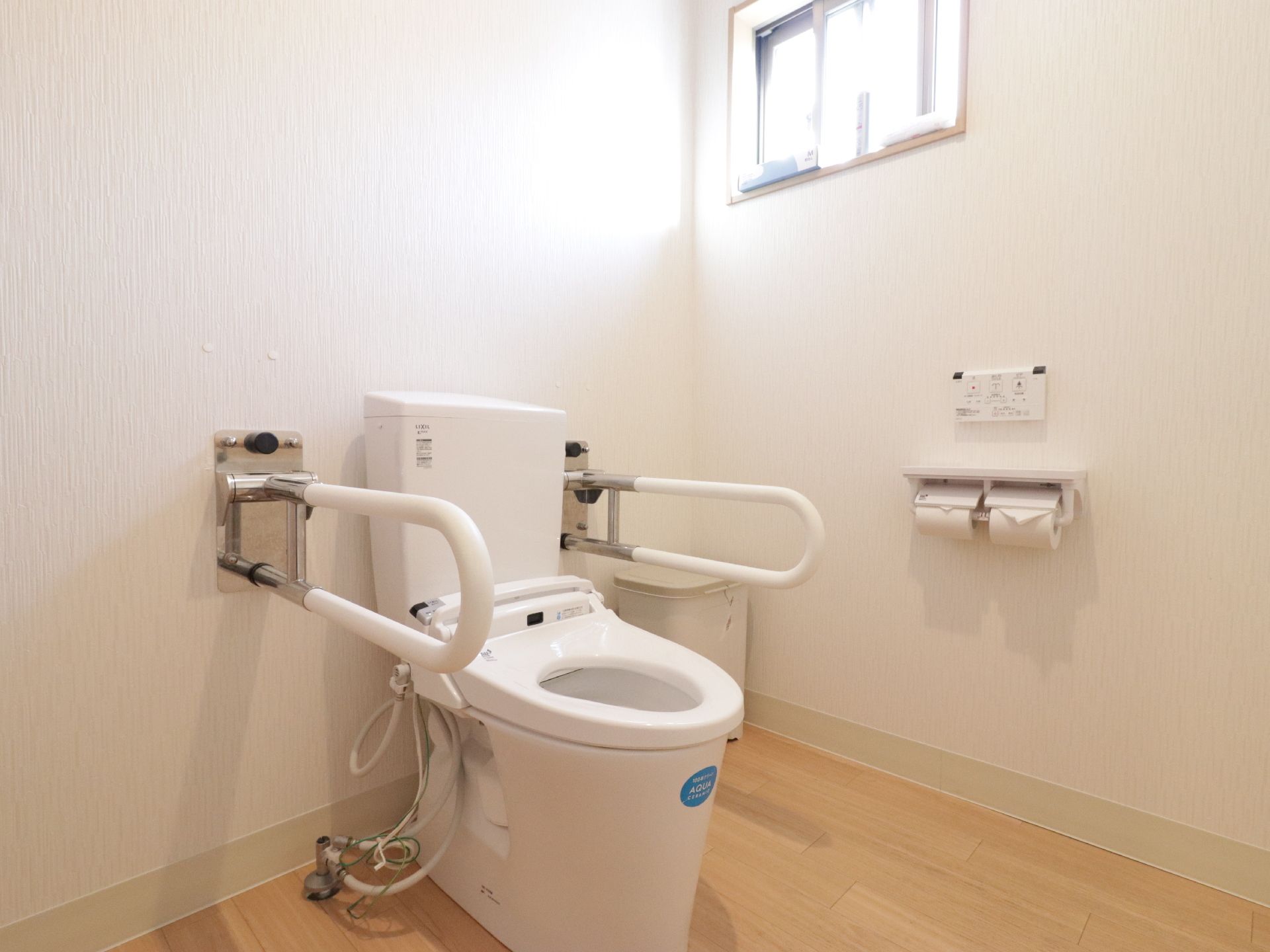 白い壁に、木目調の床が使われたトイレである。便器の両側には可動式の手すりが設置されており、車椅子のまま入る事ができる十分な広さがある。