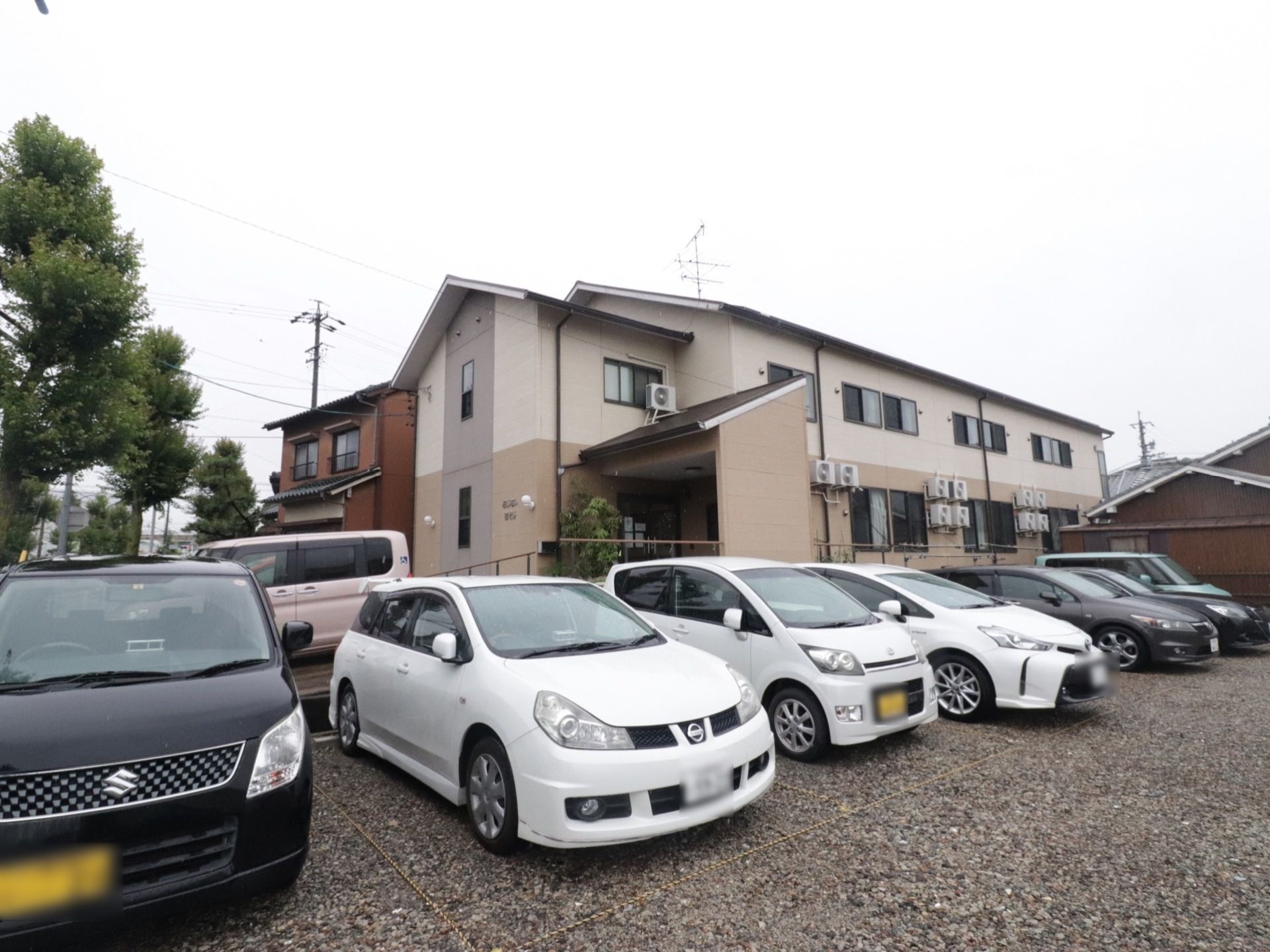 2階建ての建物があり、多くの室外機が設置されている。施設前と右側に駐車スペースがあり、多くの車が停められている。