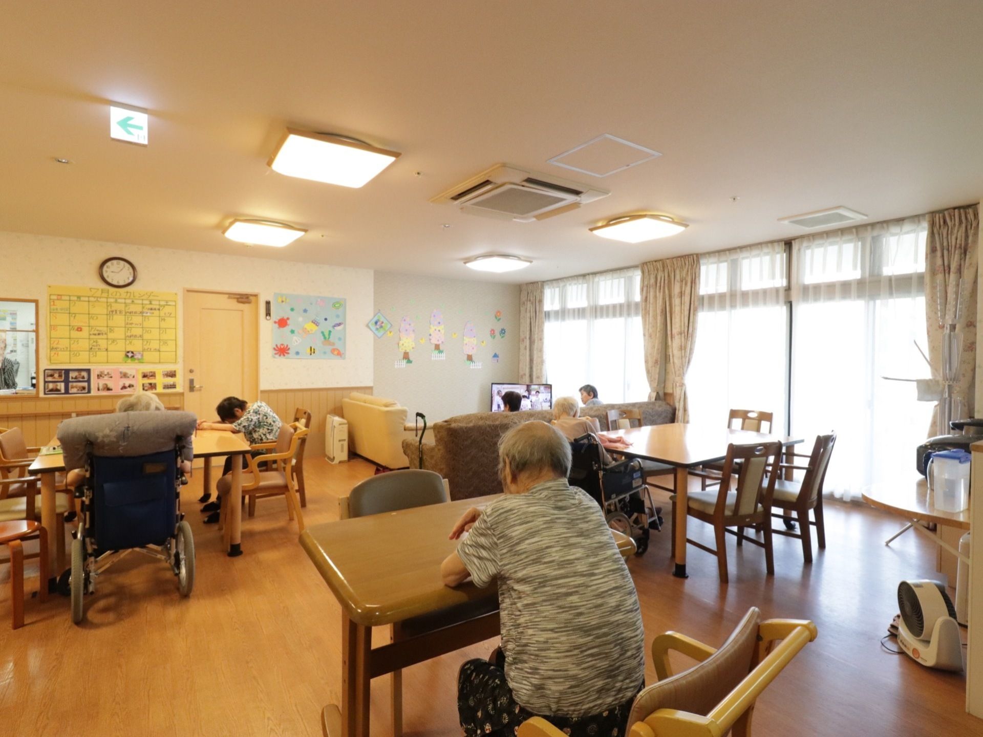 大小のテーブルセットが配置されており、入居者様が思うままに時間を過ごしている。壁一面窓が備えつけられている。