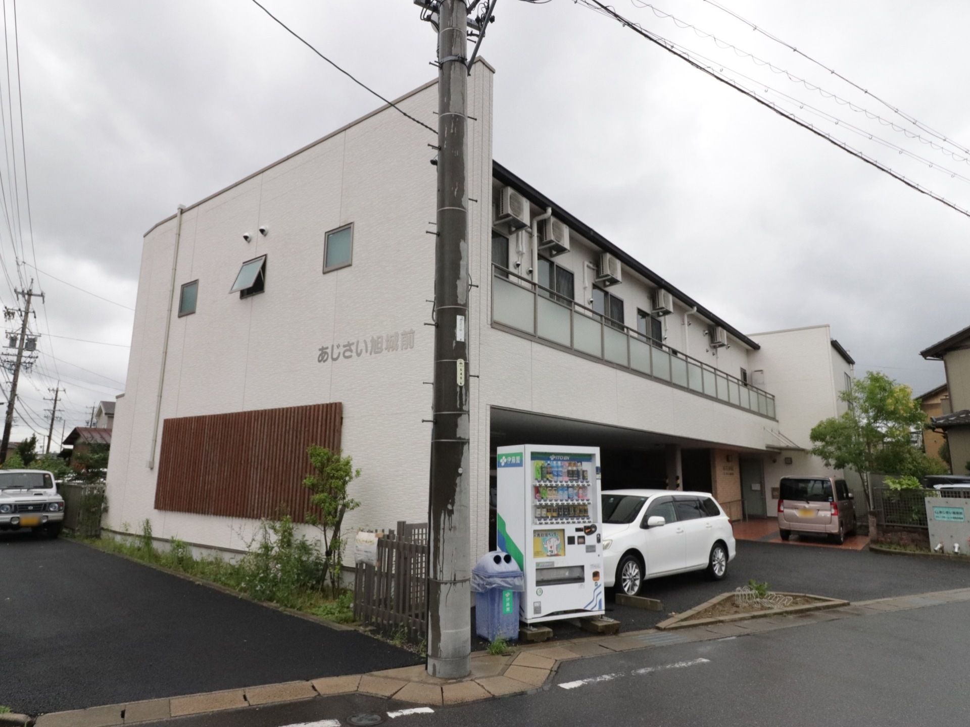 道路に面した場所に建つ、2階建ての施設である。電柱のとなりに白色の自動販売機、空き缶入れが設置されている。