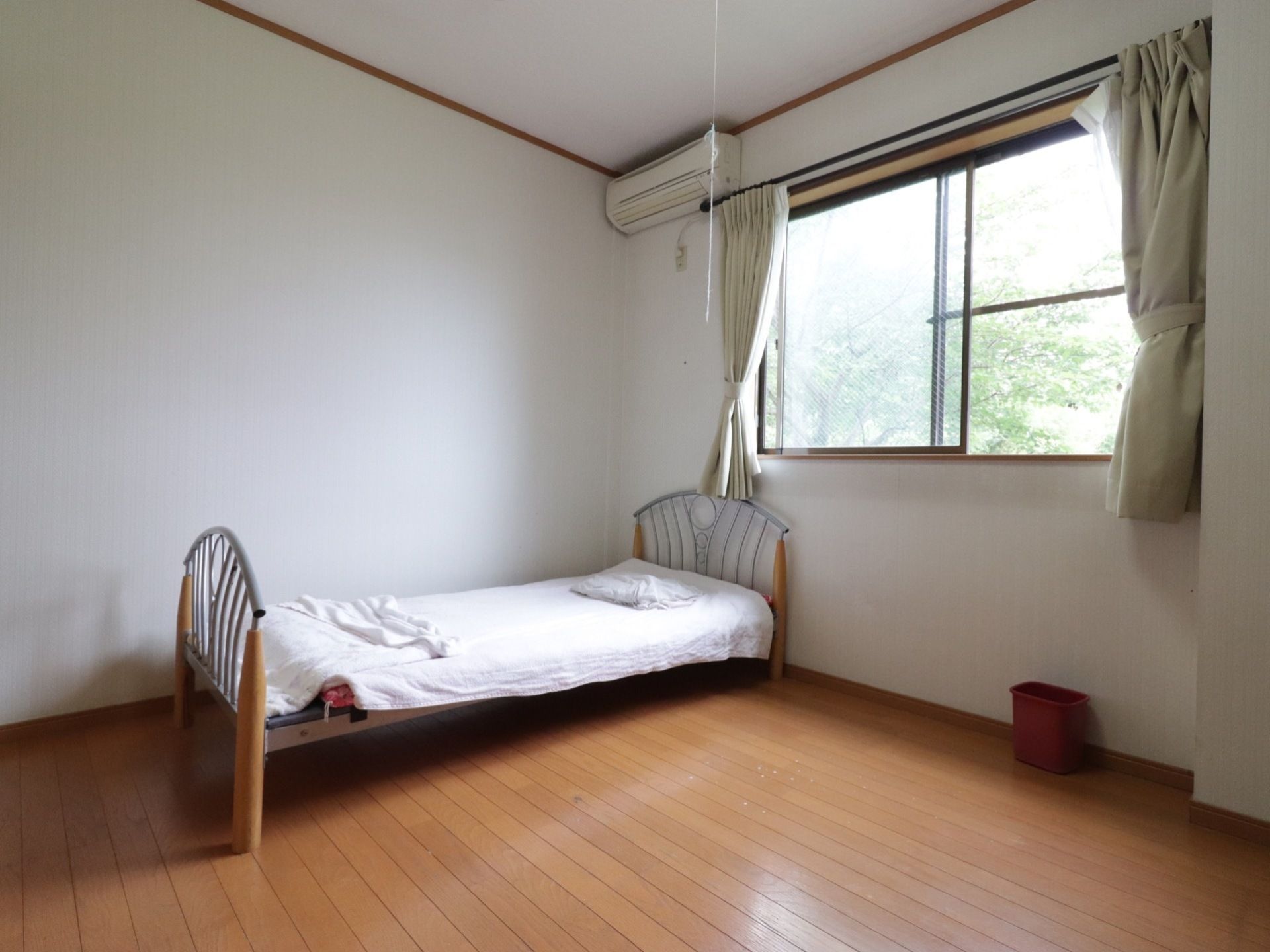 白地の壁と茶色のフローリングが組み合わさった落ち着きのある居室である。室内には縦に長いベッドのほか、エアコンが備え付けられている。