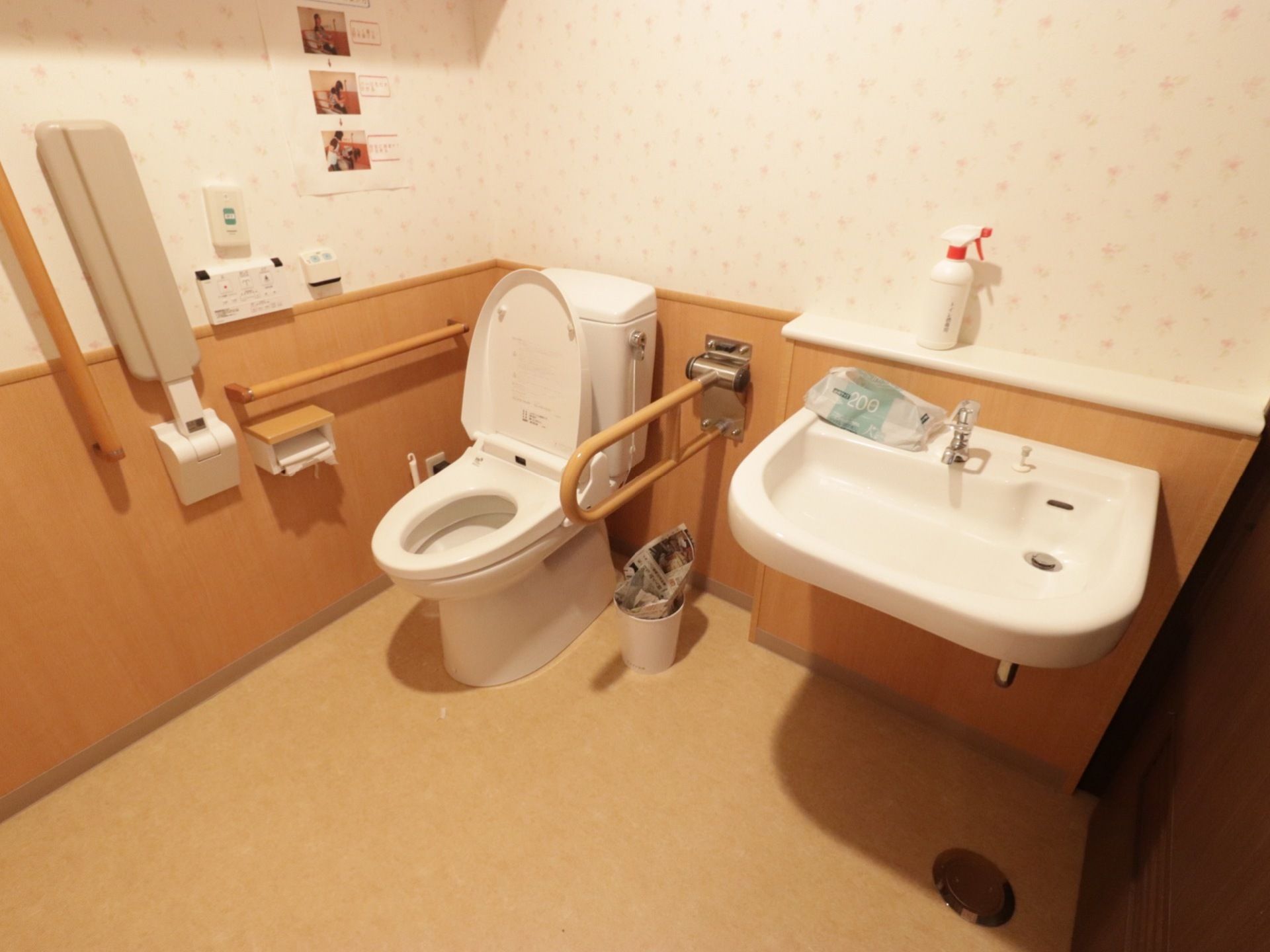 洗面台が備わった広いトイレである。可動式や水平型手すり、折り畳み式アームレストが壁に設置されている。