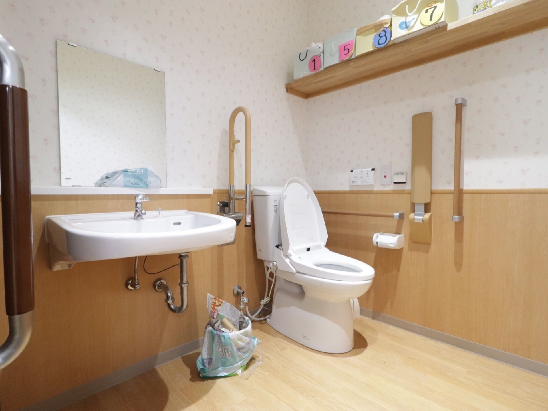 便器と洗面台が隣り合ったトイレであり、洗面台の手前には鏡が取りつけられている。便器の周囲の壁には手すりやつかまり用の棒が備わっている。