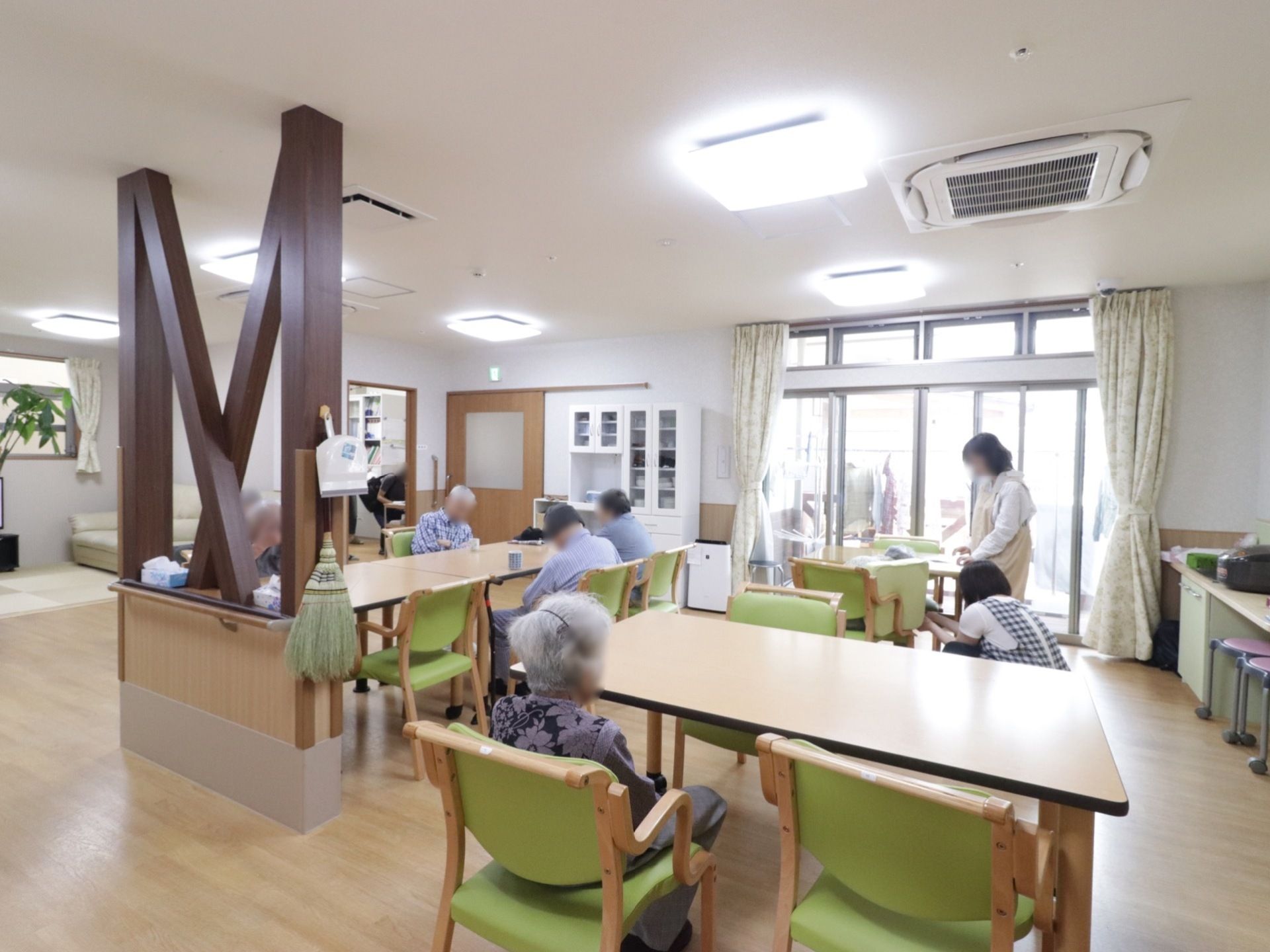 複数の長方形のテーブルが室内の各所に配置されており、それぞれの周囲に椅子が並べられている。入居者様方が椅子に腰かけ、お休みになられている。