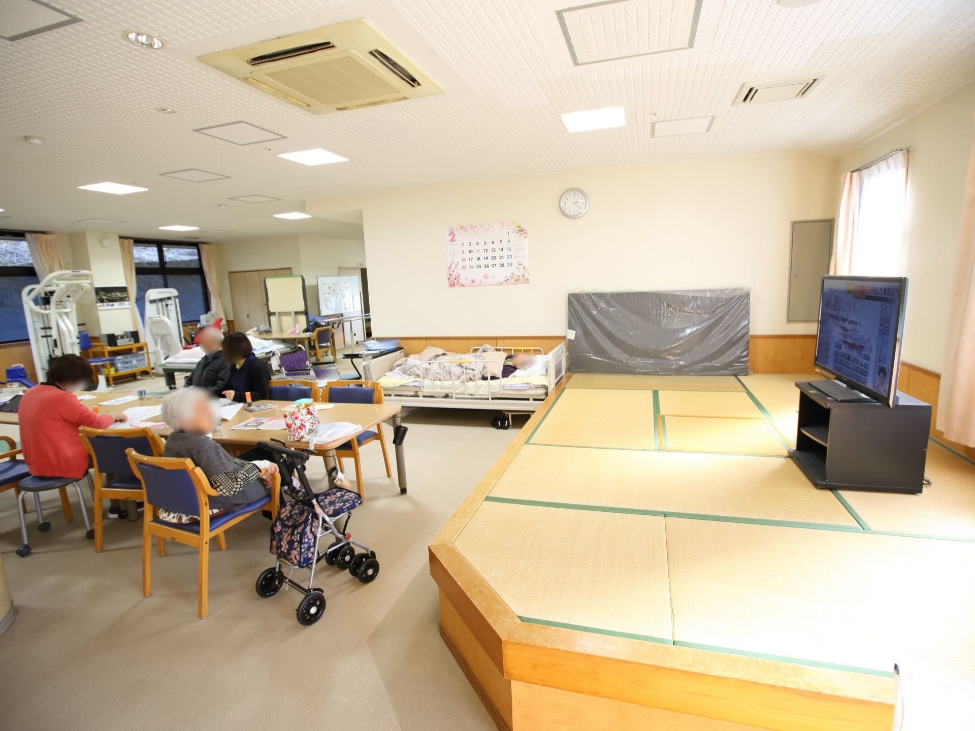 窓側に健康器具が置かれており、中央にはテーブルが置かれており、数名の入居者様が座っている様子である。
