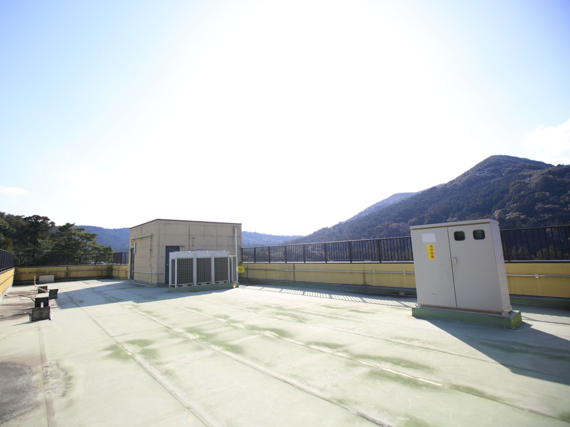周囲を一周、柵で囲まれている屋上の様子である。柵の下の壁には、手すりがある。また、建物の奥には山が見える。