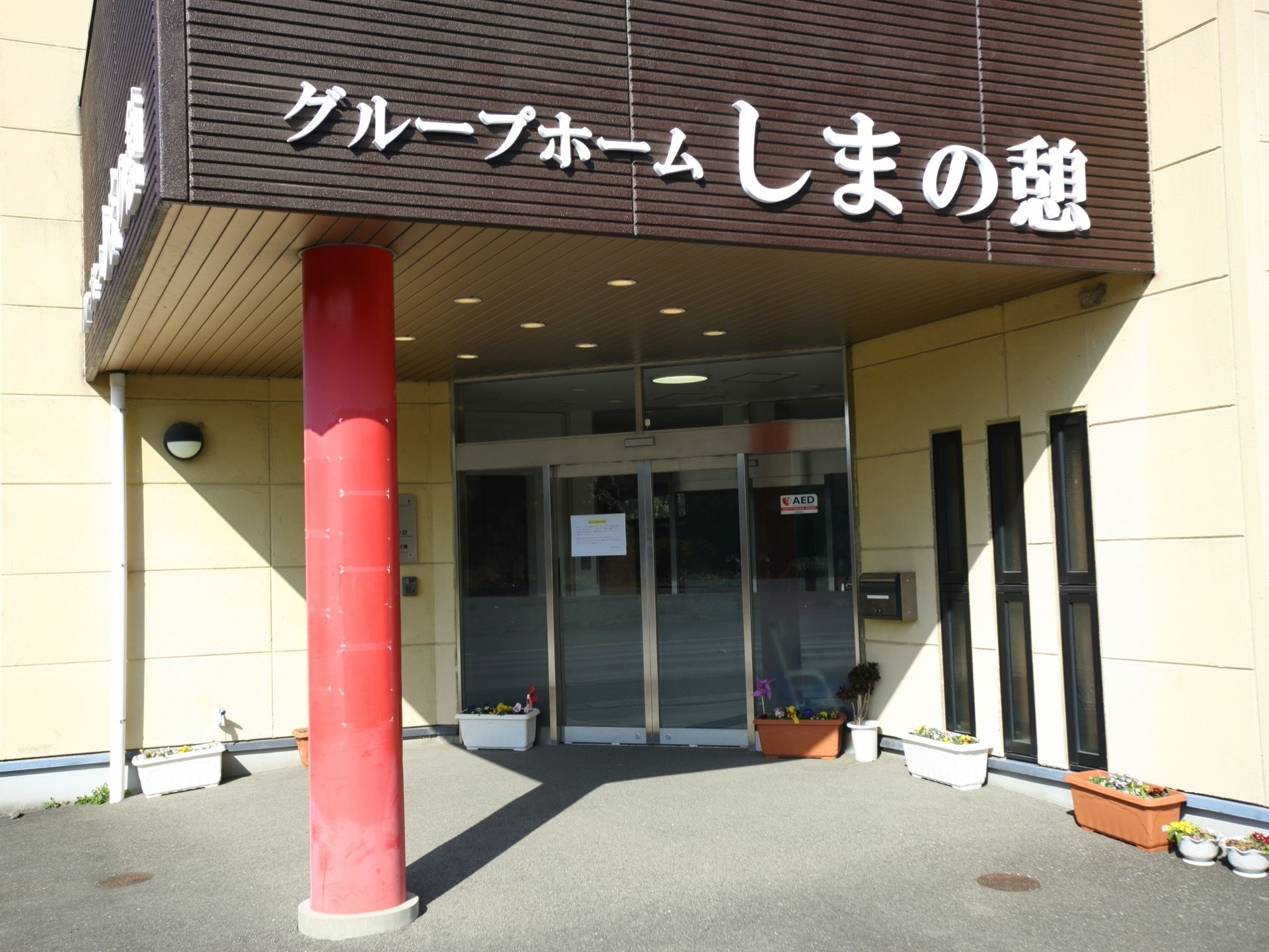 玄関前に小屋根のある入口である。赤い太い柱が１本設けられている。小屋根の側面に施設名が取り付けてある。壁には外灯が設置されている。