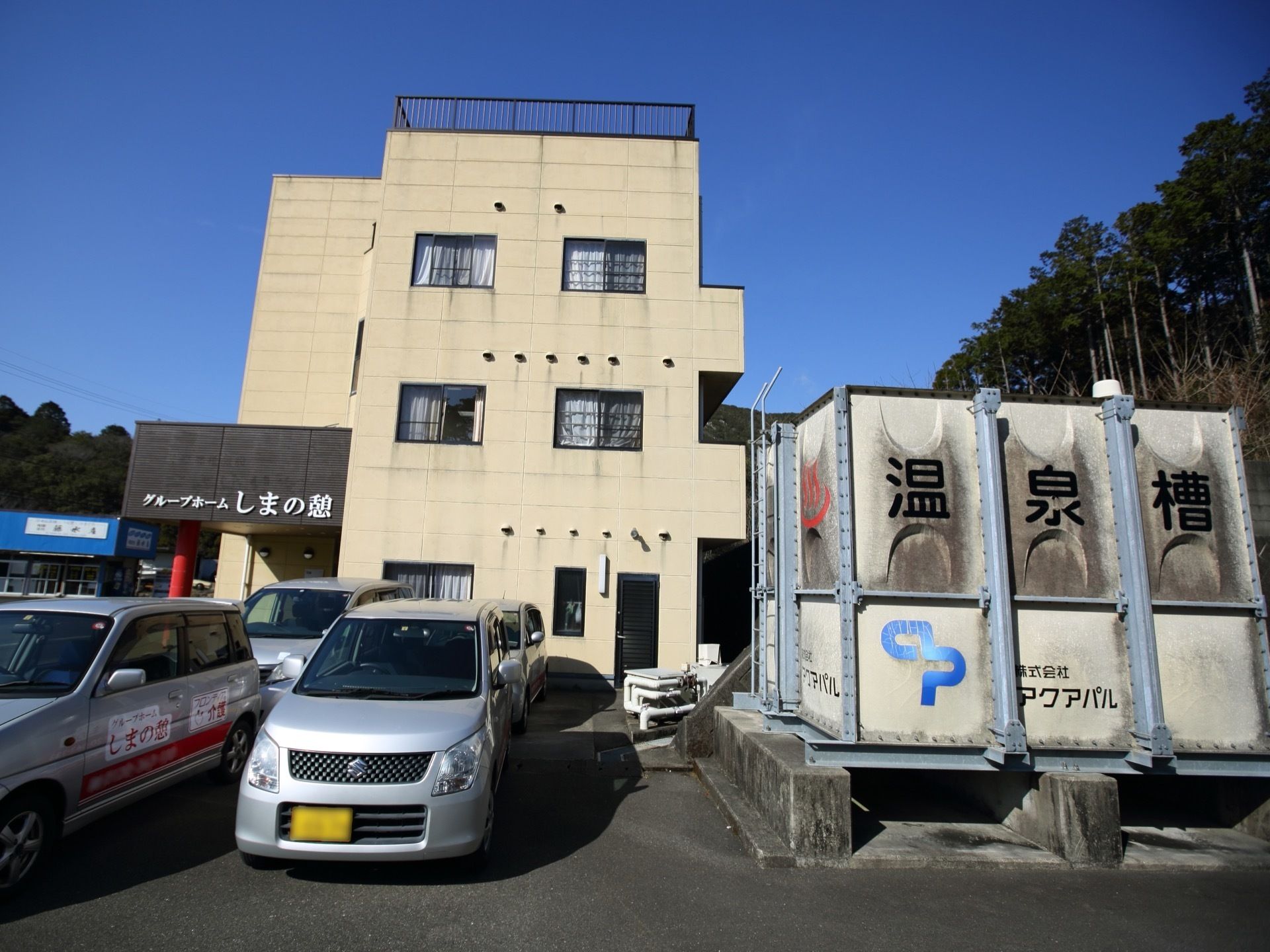 青空の下に３階建ての建物がある。建物の前には複数台の車が停まっており、温泉槽が駐車場に設置されている。