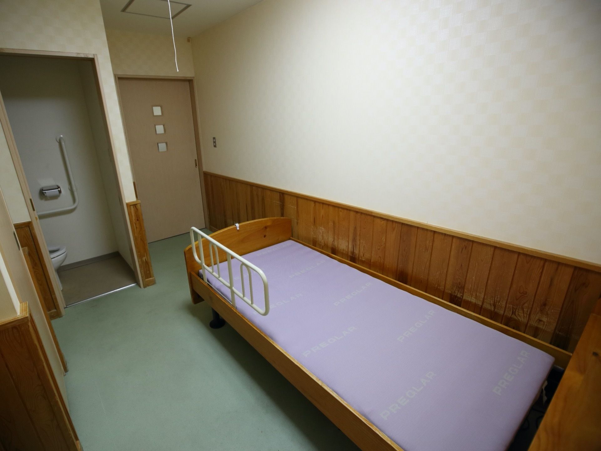 トイレの扉が開いた状態になっており、壁際にベッドが１台置かれている。天井方白い紐が１本垂れ下がっている。