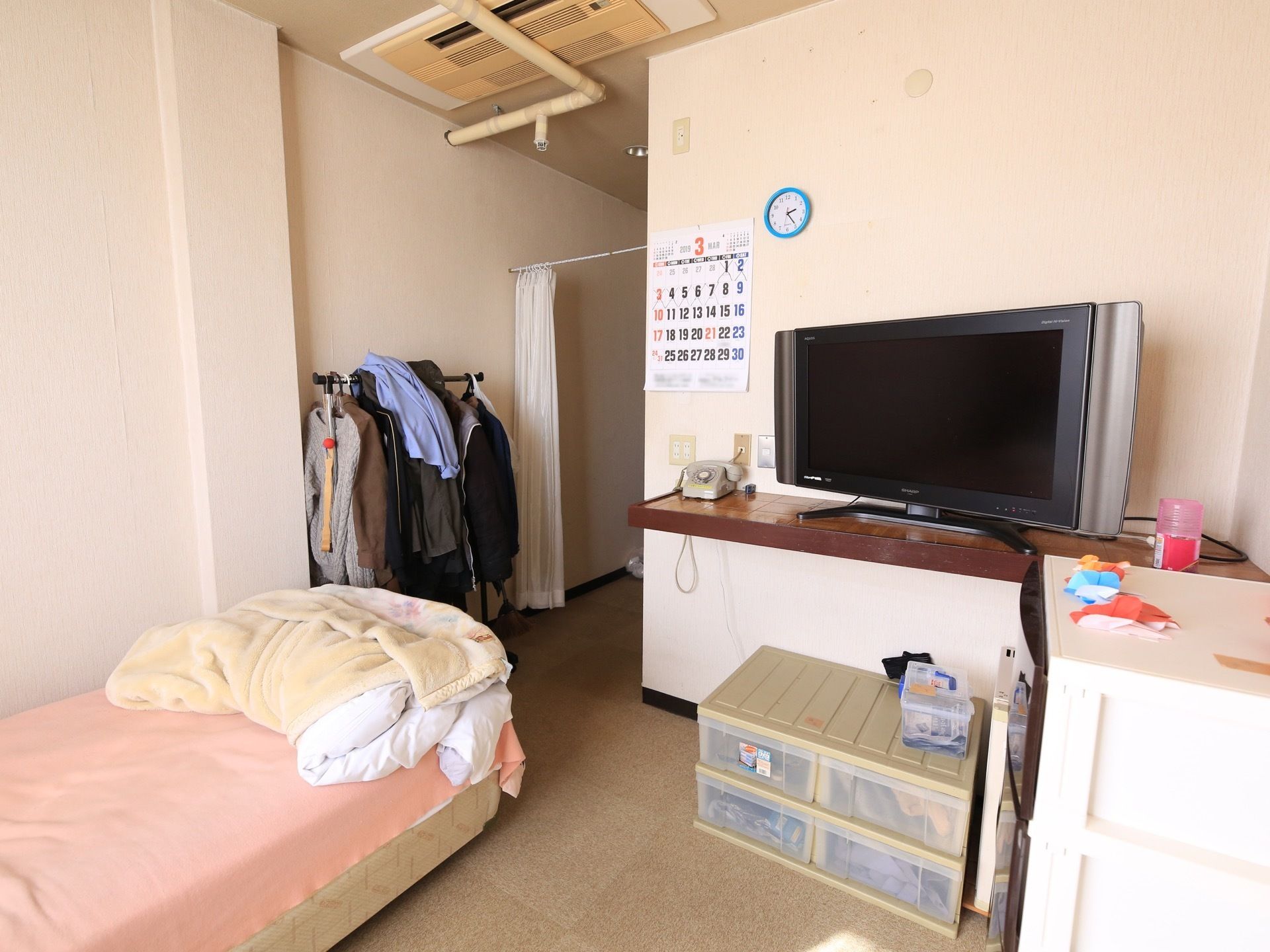 壁にカレンダーや時計が掛けられている。台の上にテレビや電話機が並べて置かれている。複数の収納ケースが設置されている。