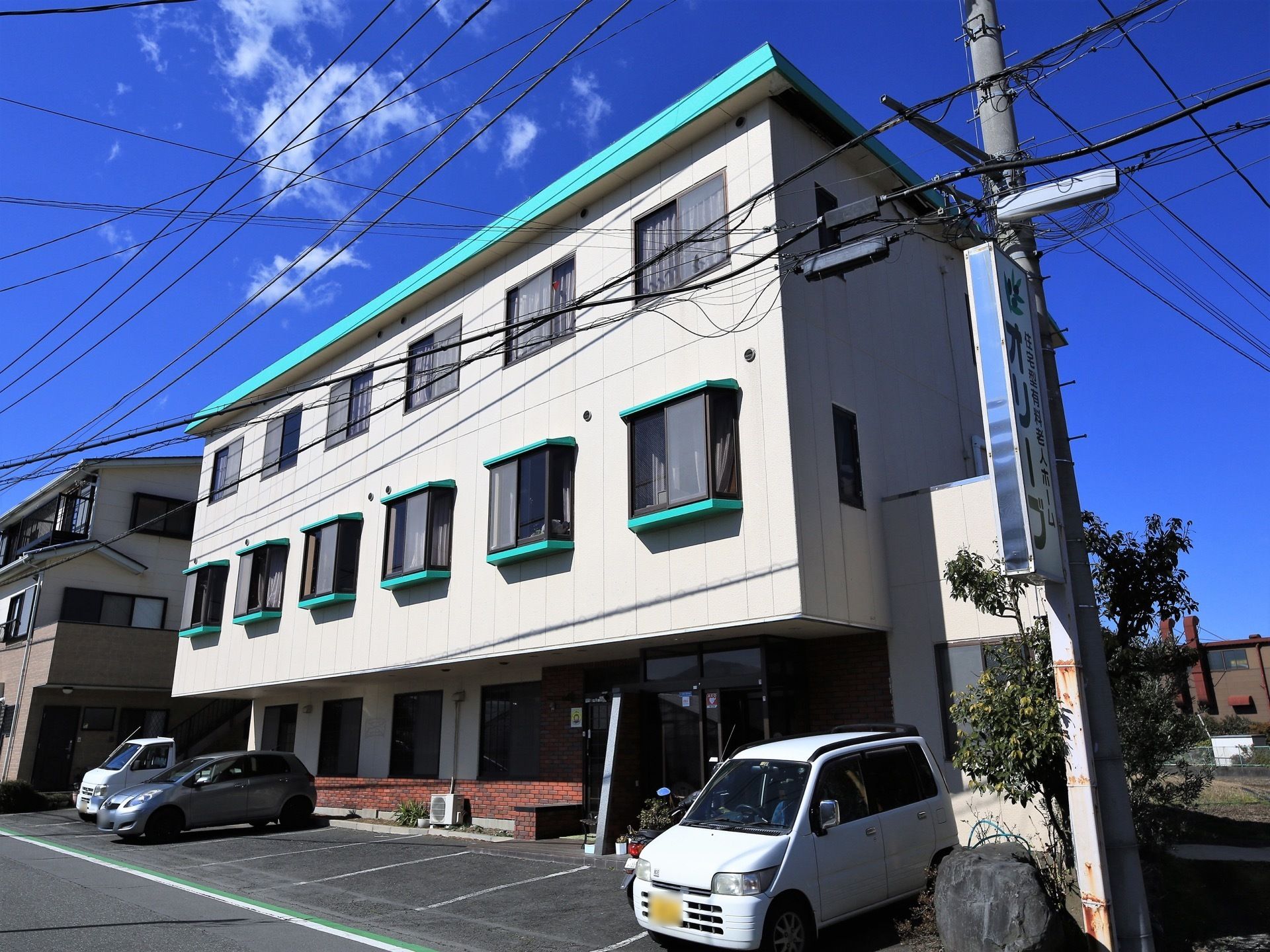 白い外壁の建物で、近くの電柱に施設名が書かれた看板が取り付けられている。施設前に複数台の車が停められている。