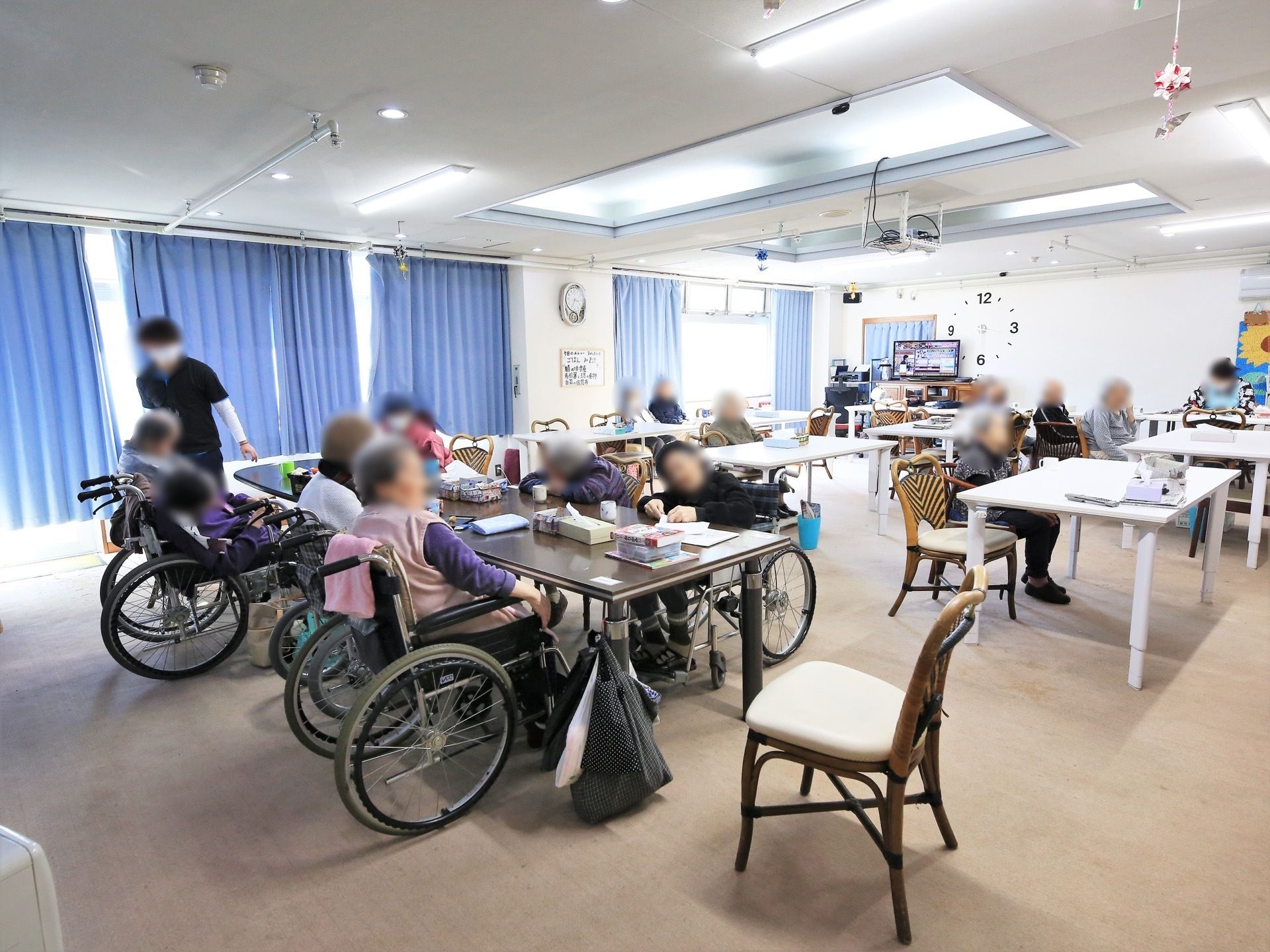 白やこげ茶色のテーブルが並べられており、入居者様たちが席についている。窓にはブルーのカーテンが掛けられている。