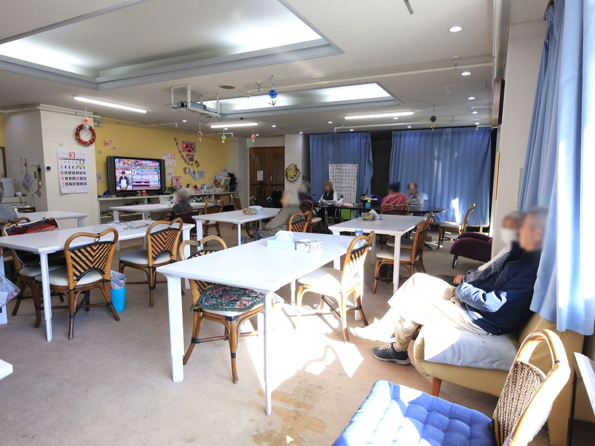 椅子やテーブルが用意されており、壁際にあるソファには入居者様が座っている。奥にはテレビが置かれていて、壁にはカレンダーが掛けられている。