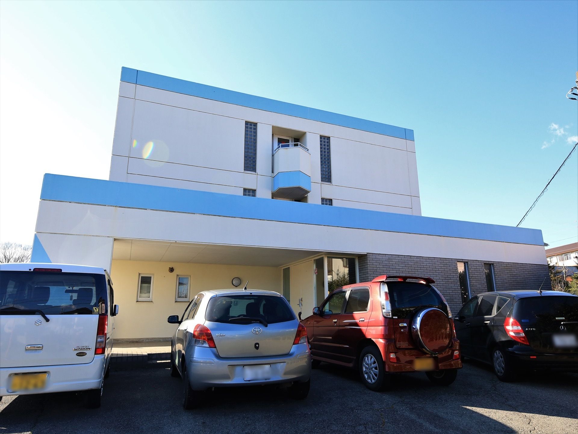 白とブルーの外壁の建物で、１階の右側はレンガ調になっている。玄関の前には駐車スペースがあり、車が数台停まっている。