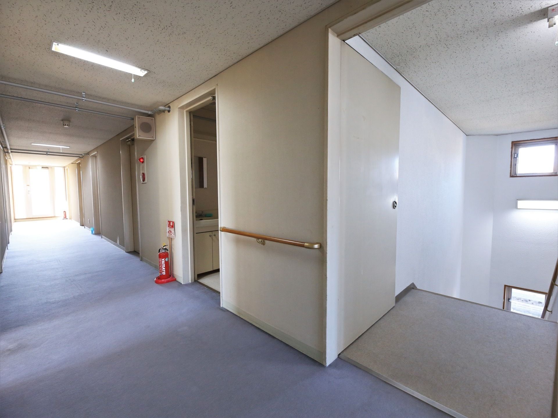 広めのスペースの通路になっていて、右側には階段がある。壁には手すりが設置され、床には消火器が置いてある。