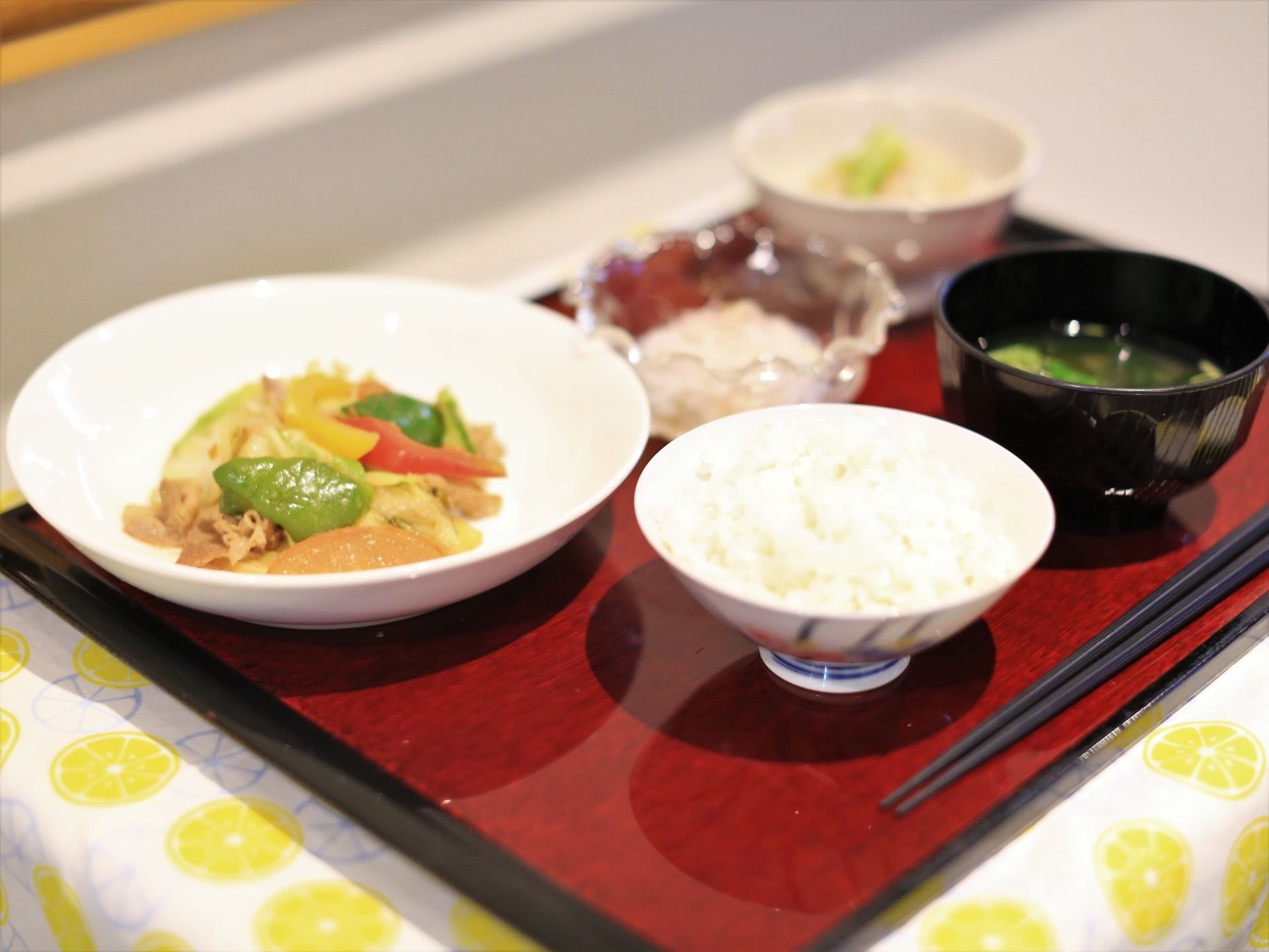 ごはんや味噌汁の他に、白い皿に入った野菜炒めや、小鉢などが置かれている。黒いお箸も添えられ、トレーの下にはテーブルクロスが敷かれている。