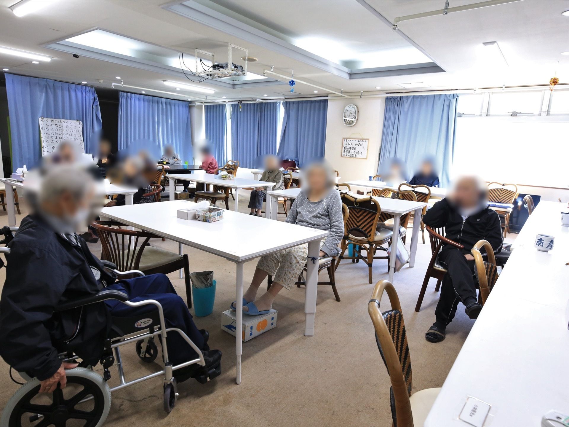 白いテーブルが複数置かれており、入居者様が椅子に座っている。壁には時計が掛けられており、窓にはブルーのカーテンが取り付けられている。