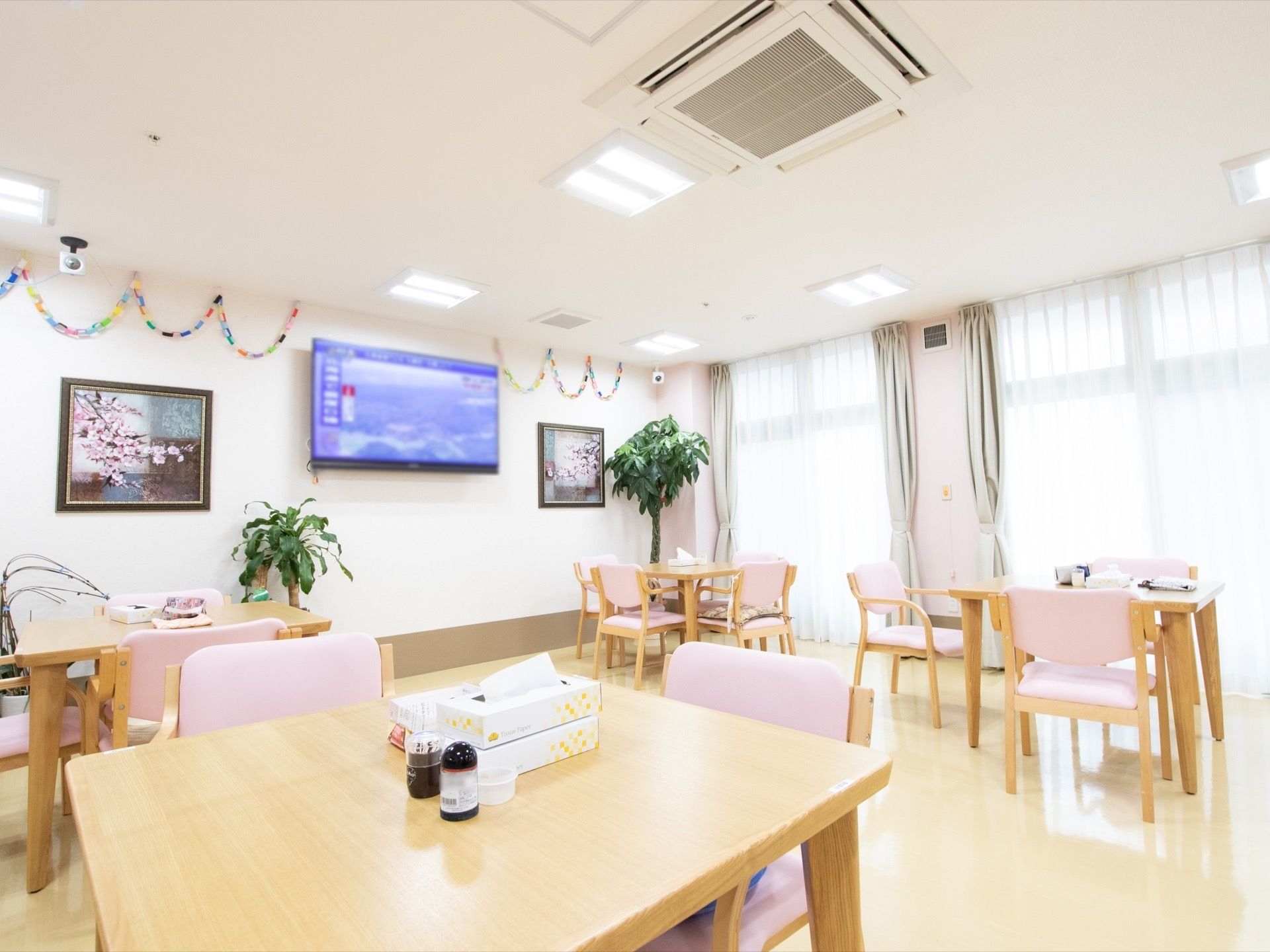 窓が大きく日差しがたっぷり差し込む。壁には画面の大きな液晶テレビと桜の写真が掛かっている。観葉植物が置いてある。
