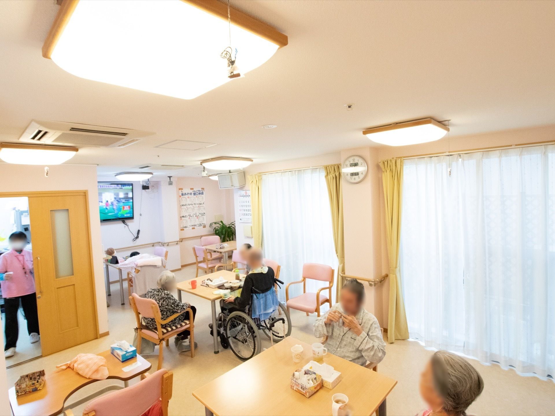 共有スペースに入居者様が集まっている。大きな窓や天井の大きな照明で明るい。スタッフが入居者様のお世話をしている。