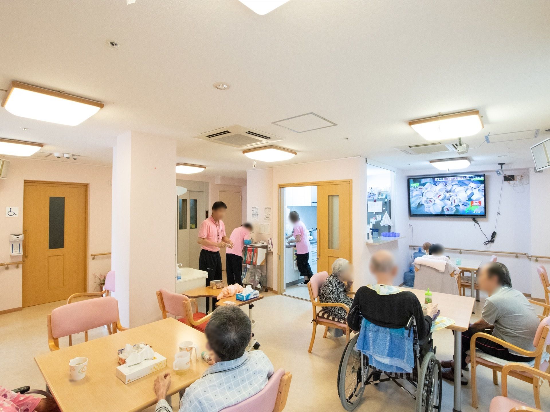 共有スペースに入居者様が集まっている。スタッフが３名、洗い物や記録などを行っている。大きな照明がいくつもある。