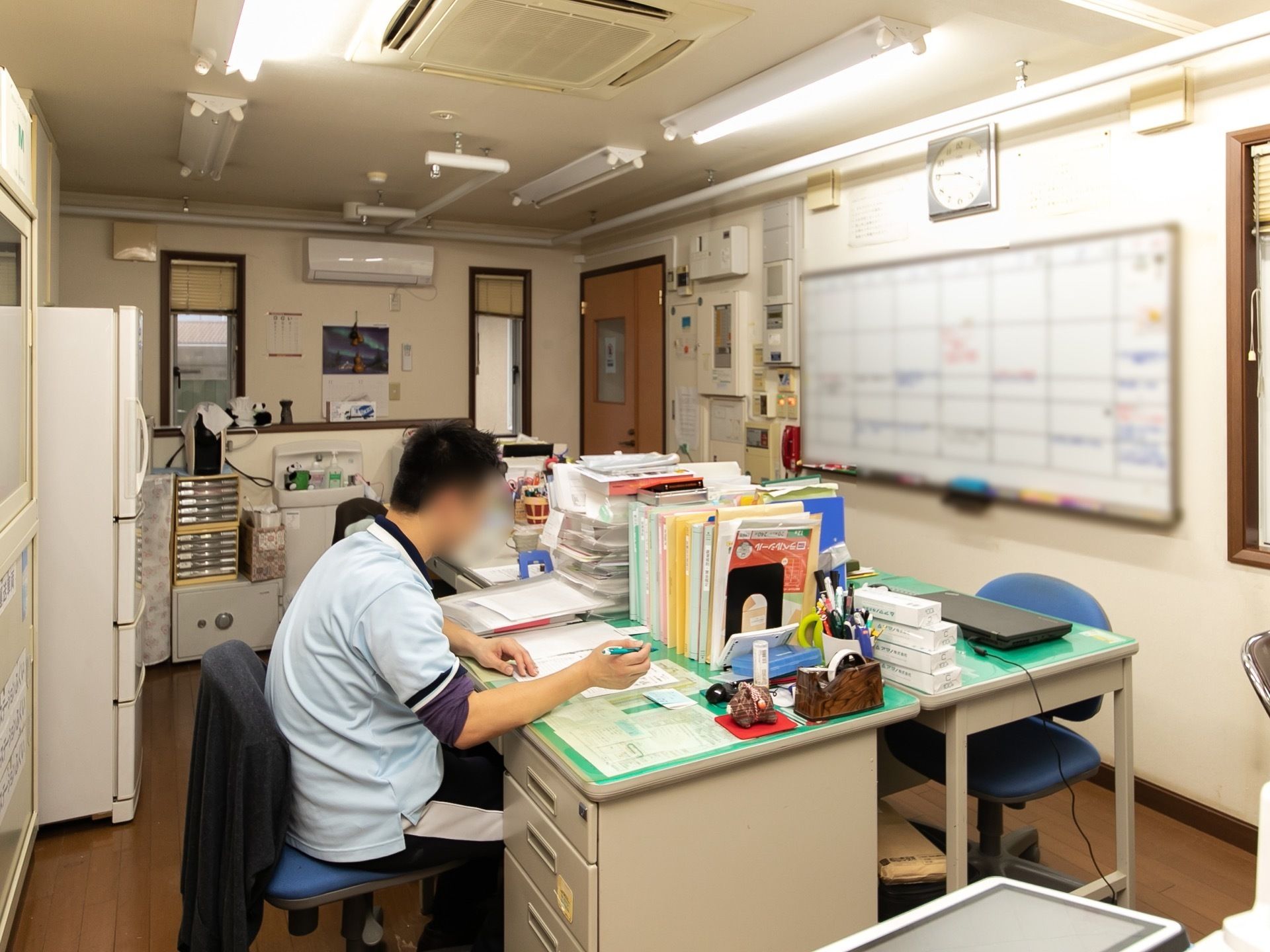 整理整頓が行き届いた事務室で、スタッフがデスクに座り作業をしている。壁にはホワイトボードが設置されている。