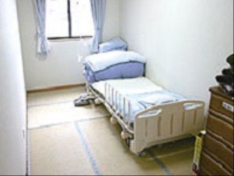 畳の部屋で、畳の上に介護用のベッドが用意されている。ベッドの側にはタンスが置いてあり、その上にも物がある。