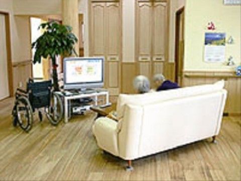 白いソファには入居者様が２人並んで座り、テレビを眺めている。ソファの近くの壁にはカレンダーがかけてある。