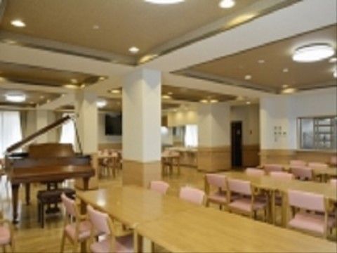 広々とした食堂に、茶色のグランドピアノがある。テーブルや椅子がきれいに並べられている。椅子の背当てと座面はピンク色である。