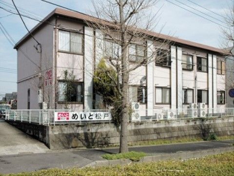 施設は２階建ての建物で、ベージュ色の外壁である。建物奥が駐車場になっている。道路側の柵に、施設名が書かれた看板が取り付けられている。