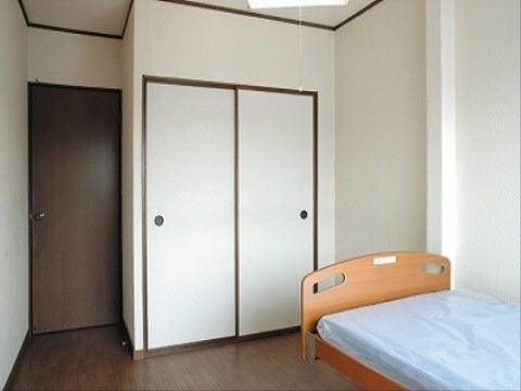 白い壁に焦げ茶色のドアがある居室である。収納スペースの扉も焦げ茶色で縁取られており落ち着いた印象になっている。ベッドが備えてある。