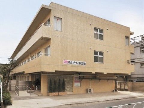 ベージュ色の３階建てのマンション風の建物。エントランス周りには、木々が植えられ、入口にはスロープが設置されている。