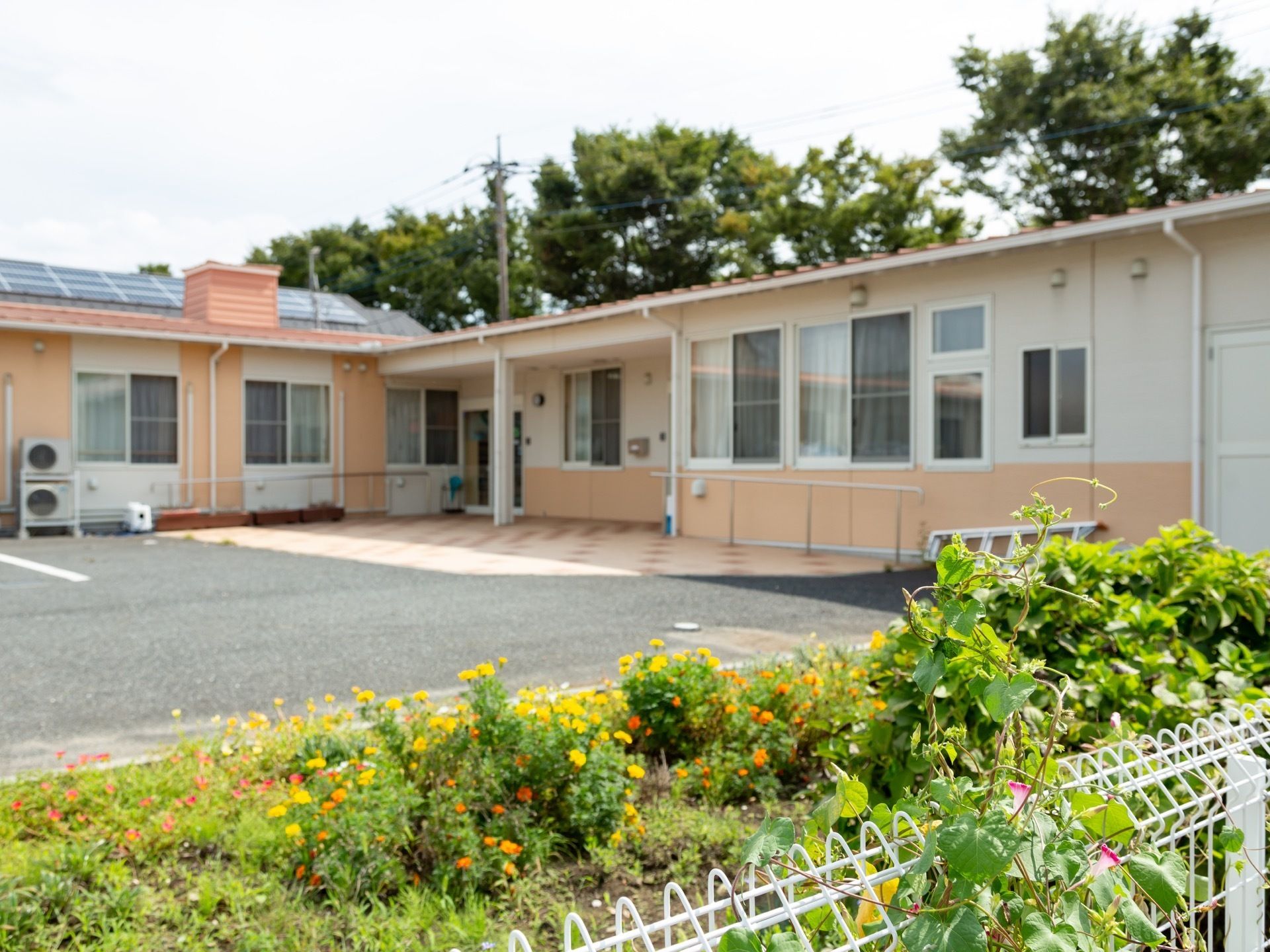 白色と薄いオレンジ色の平屋建ての建物がある。敷地内の花壇に黄色やオレンジ色の植物が植えられ、白いフェンスが設置されている。