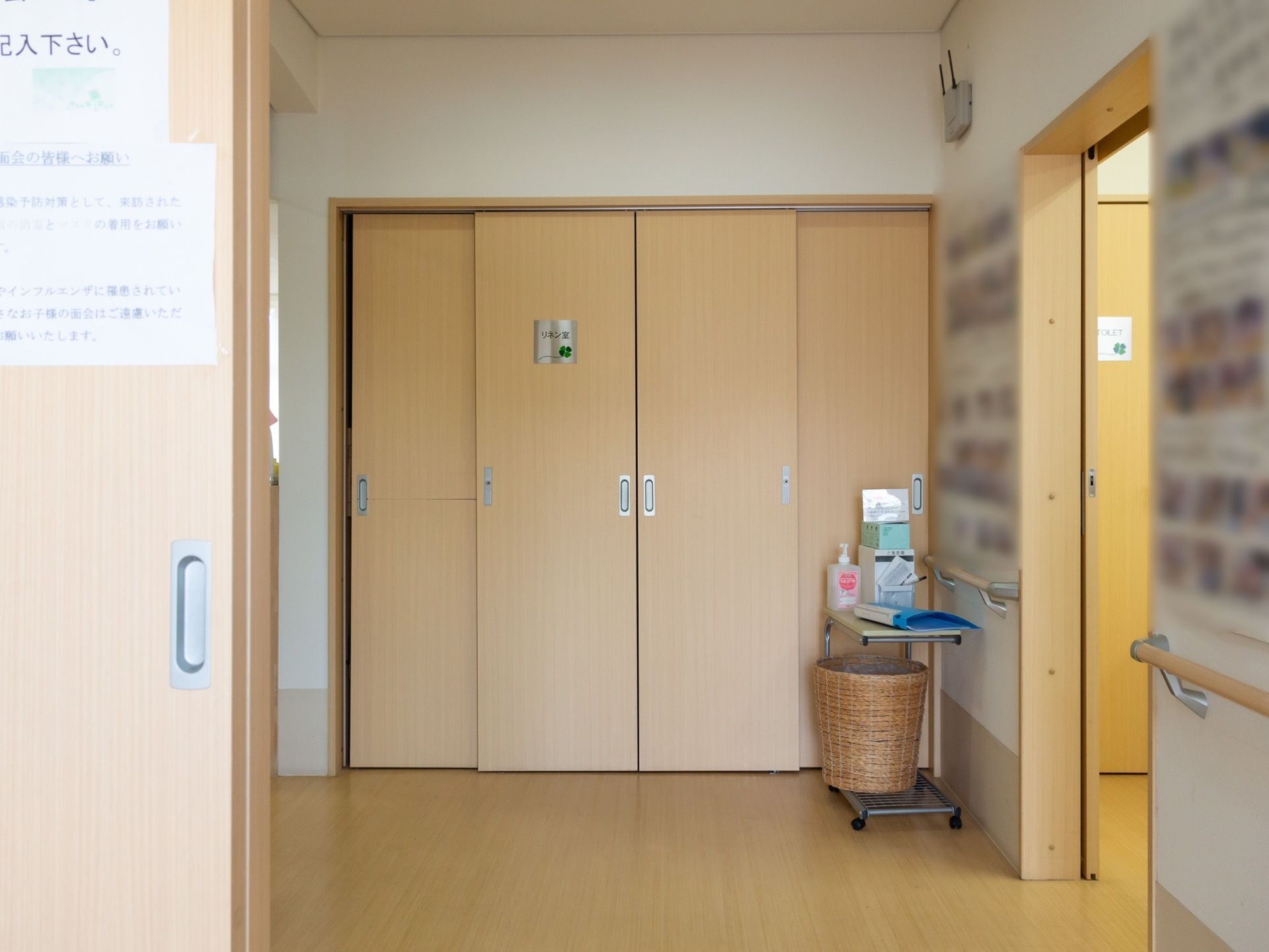 リネン室と表示されたパネルが貼られた４枚扉の入り口がある。扉の前にかごと消毒液が乗せられたキャスター付きの台がある。