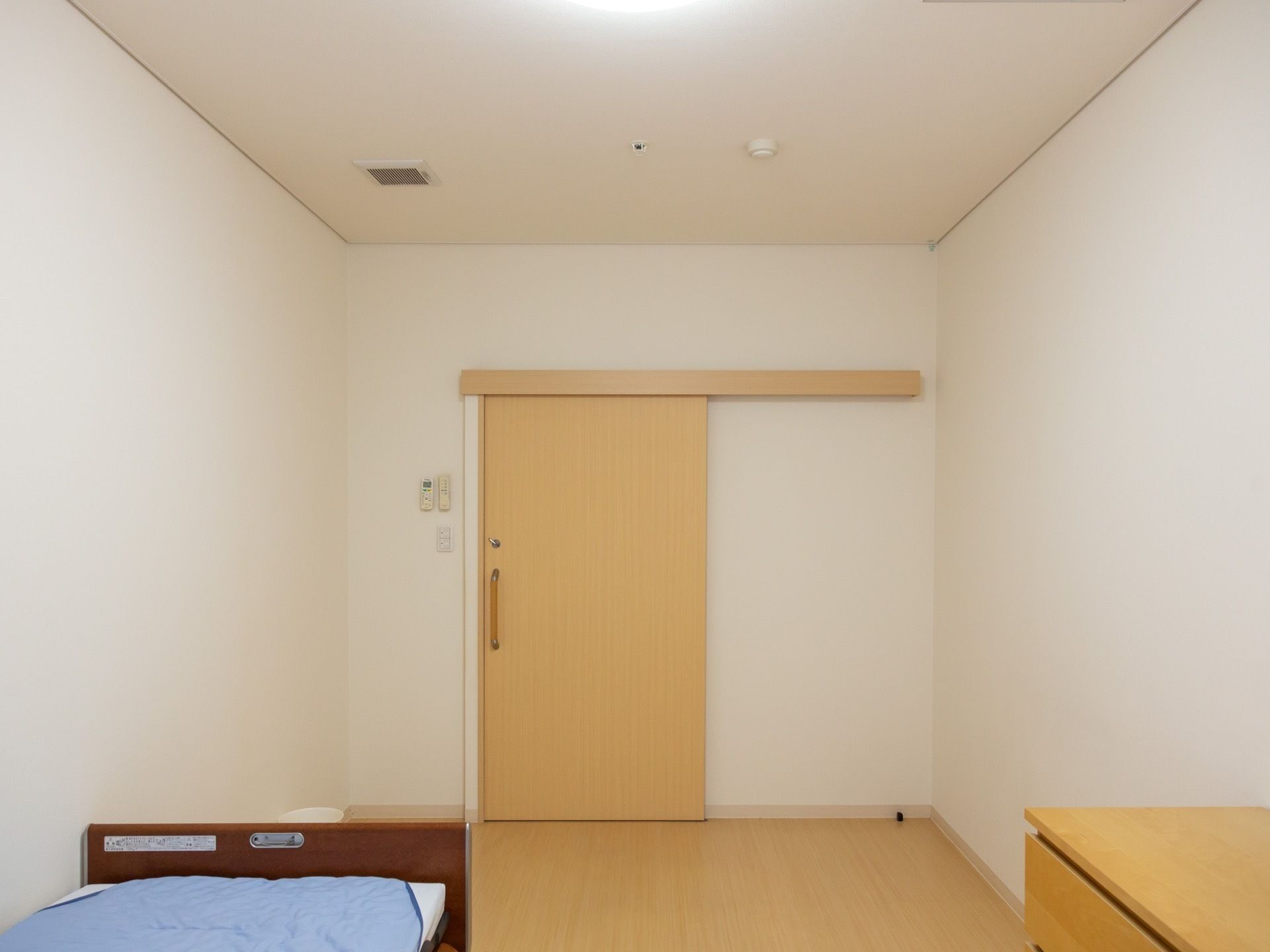白色の壁と淡い色の木目のフローリングのシンプルな部屋である。スライドドアの横の壁にエアコンのリモコンが設置されている。
