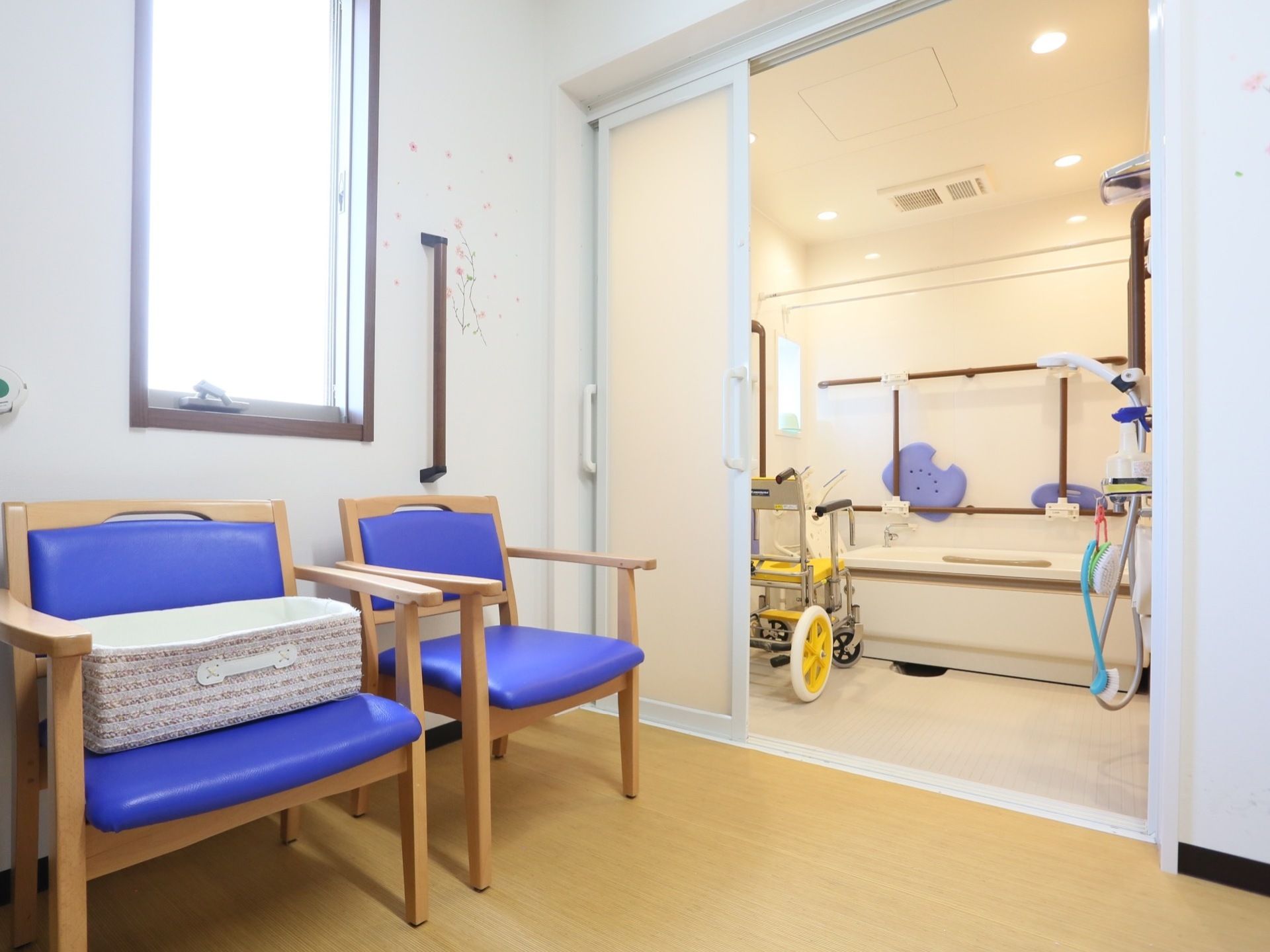 窓のある明るい脱衣所の壁には、花の絵があしらわれており、その前に椅子が２脚置かれている。奧に、横幅の広いユニットバスがある。