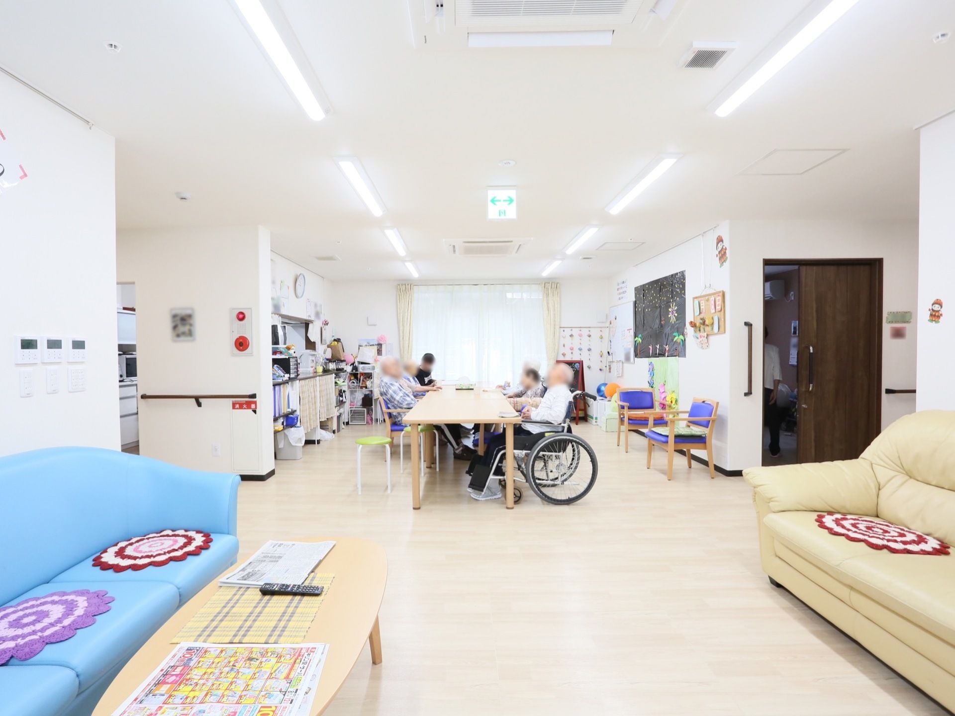 ソファが向かい合って配置されている。奧にはテーブルが一列に並べられており、椅子や車椅子に腰掛けた数人がテーブルに向かっている。
