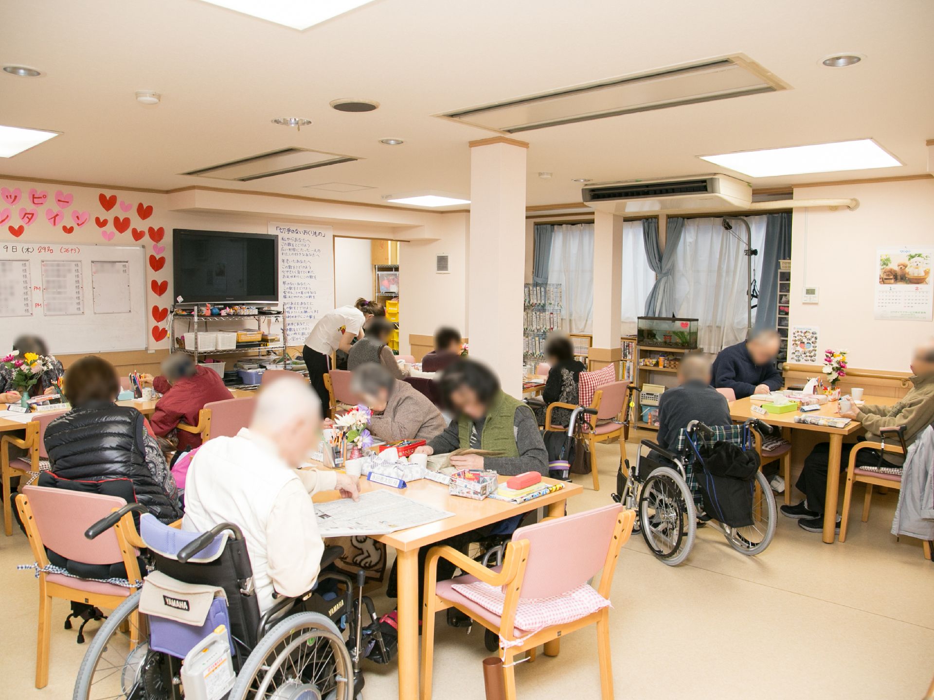 ゆったりとしたラウンジには、明るい照明が設置されている。多くの車いすのご入居者様がおり、テーブルで書き物をしている。