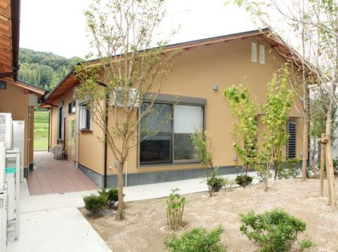 オレンジの土壁風の落ち着いた佇まいの建物である。施設前に植栽を植え、背後には山々の緑が広がっている。