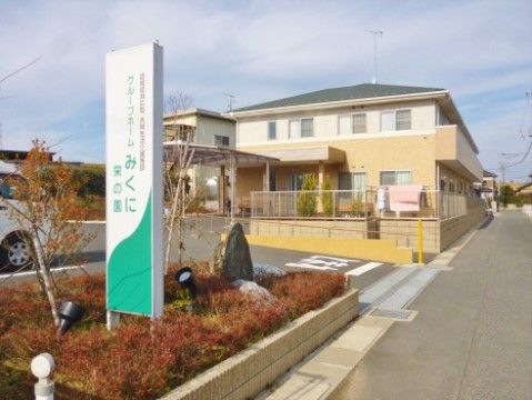道路沿いに低い塀に囲まれた、民家のような２階建ての建物がある。手前の花壇には施設名の看板が設置され、駐車場へも入りやすい立地になっている。