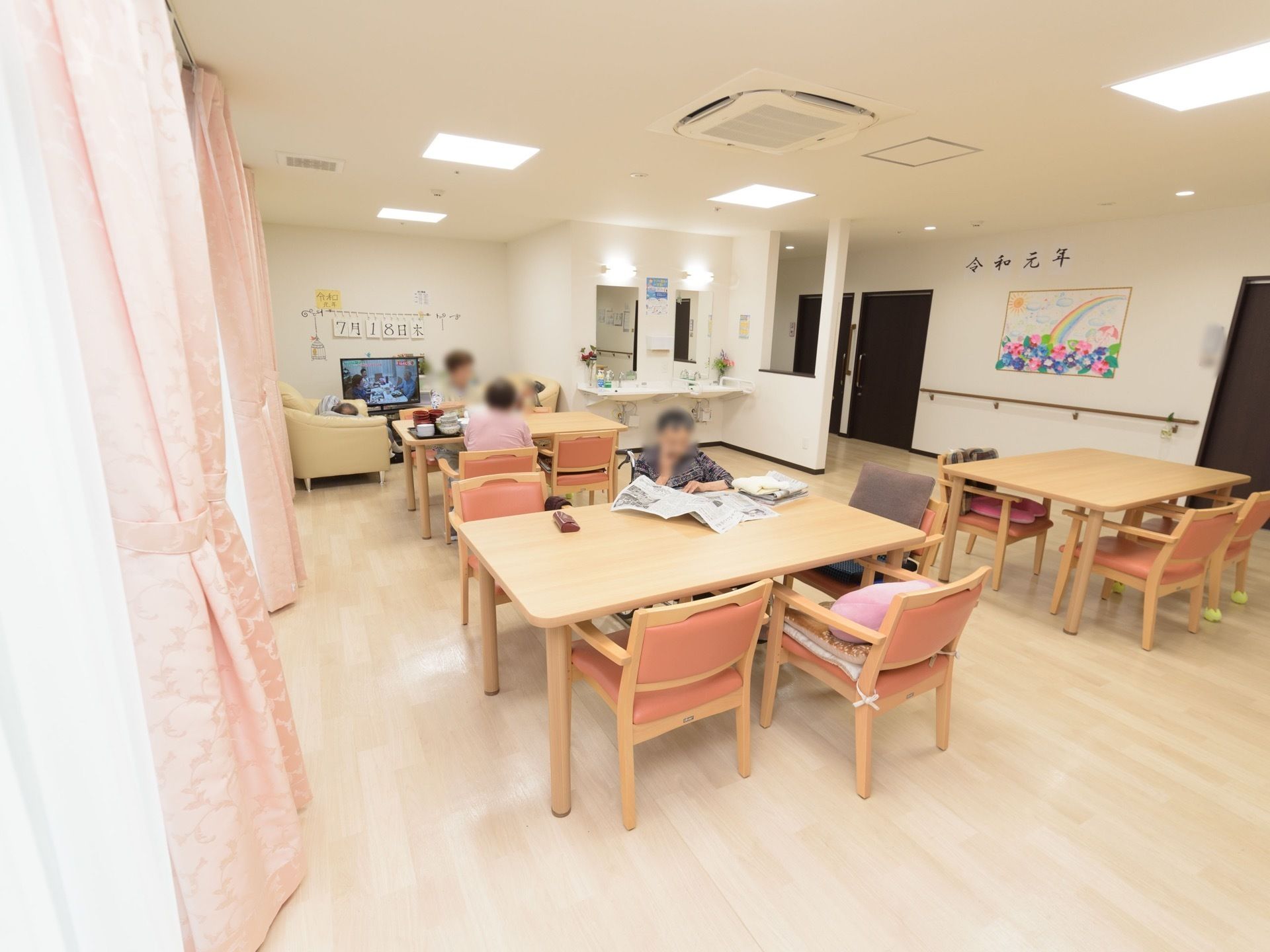 洗面台、テレビ、ソファ、ダイニングテーブルなどが設置された共有スペース。周囲の通路の壁には、折り紙細工なども飾られている。