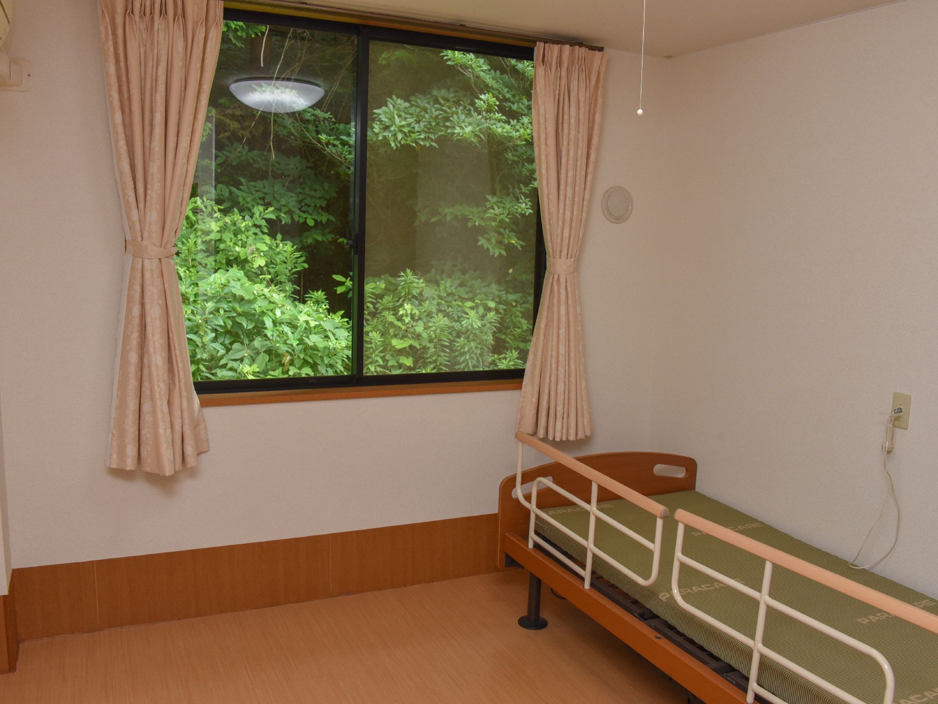 個人の居室にはベッドが置かれており、落下防止柵も設置されている。奥の壁には窓があり、外には緑の木々を見ることができる。