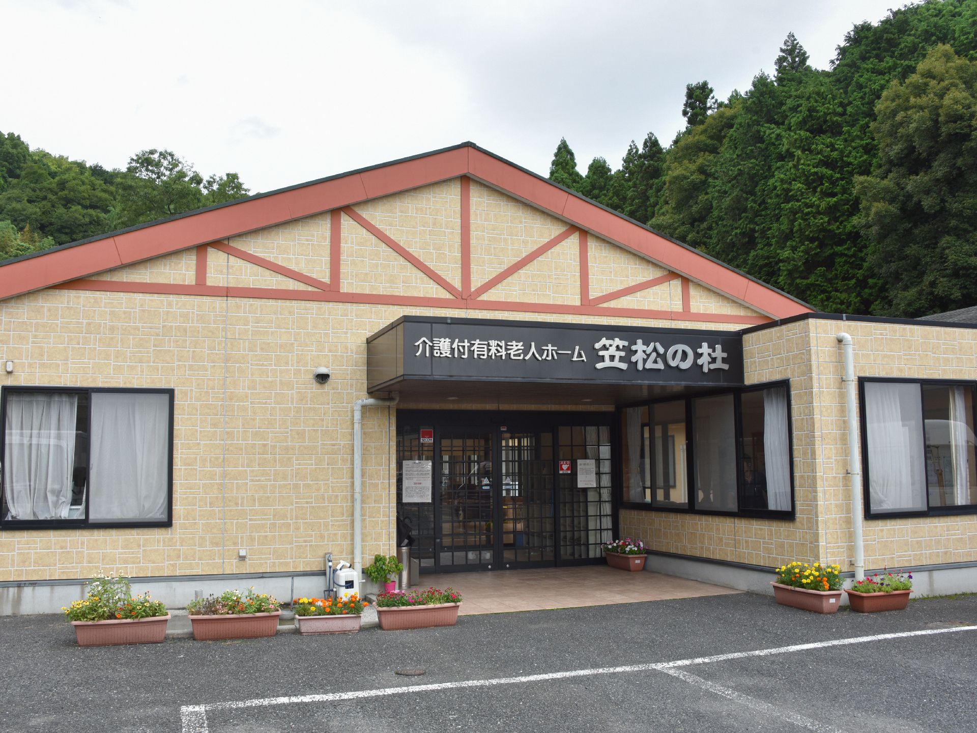 三角の屋根が特徴的な施設の外観。自動ドアの上には大きく施設の名前が記されている。建物の後ろは木々に囲まれている。