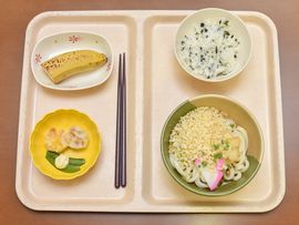2つのスペースに区切られたお盆の上には4つのお皿が置かれ、それぞれにシュウマイやうどん、バナナなどが乗せられている。